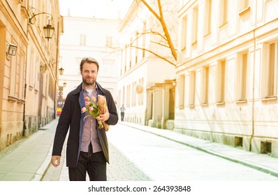 Happy Smiling Forty Years Old Caucasian Man With Baguette And Flower Bouquet Walking Down The Street. City Buildings As Background.