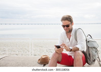 Happy smiling forty years old caucasian tourist man looking at mobile phone outdoor. Beach and sea as background - summer holiday traveling.