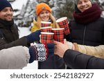 Happy smiling friends relaxing, clinking a glass of hot coffee among snow covered trees in snowy winter park outdoors. Young people in winterwear having fun in forest drinking hot beverage.