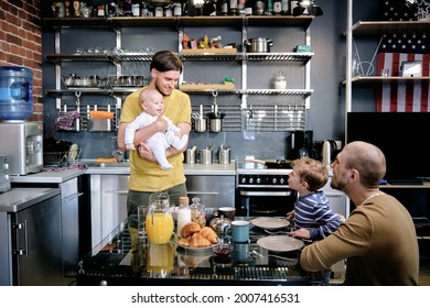 Happy Smiling Gay Man Introducing Newborn Baby To His Older Son At Family Breakfast