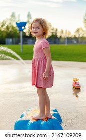 Happy Smiling Girl Playing At Splash Pad With Water. Little Beautiful Kid Standing At Playground With Fountains In Summer