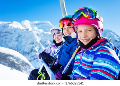 Happy Smiling Girl Sit On Chairlift Wearing Helmet And Bright Ski Outfit With Friends Over Beautiful Mountain Peak