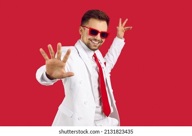 Happy smiling good looking brunet man in elegant white suit and party glasses dancing on red background. Studio shot of show presenter, TV commercial actor, singer, dancer or wedding event toastmaster - Shutterstock ID 2131823435