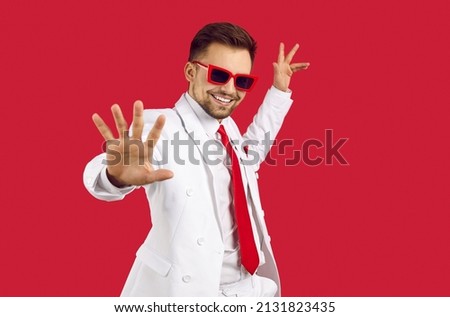 Happy smiling good looking brunet man in elegant white suit and party glasses dancing on red background. Studio shot of show presenter, TV commercial actor, singer, dancer or wedding event toastmaster