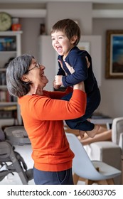 Happy Smiling Grandmother Playing With Her Little Grandson Holding Him In Her Arms In The Air. Babysitting. Home Lifestyle. Different Ages Family Bonding.