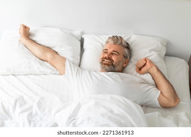 Happy smiling handsome middle aged man lying in bed alone, stretching arms after waking up, feeling well-rested, ready for new day, looking at copy space, view above - Powered by Shutterstock