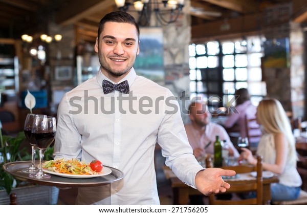 レストランでサラダとワインの入ったグラスを持つトレーを持つレストランで 笑顔のハンサムなウェイター の写真素材 今すぐ編集