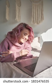 Happy Smiling Hipster Gen Z Woman Student With Pink Hair Wearing Hoodie Using Laptop Computer Tech Device Relaxing Lying In Bed At Home Surfing Internet Learning Online On Pc In Bedroom. Vertical