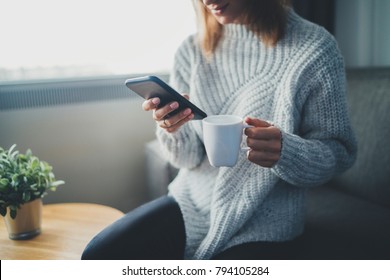 Happy Smiling Hipster Girl Using Smartphone Device While Chilling At Home Wearing Cozy Knitted Sweater, Attractive Young Woman Chatting With Friends At Social Network While Sitting On Sofa At Home