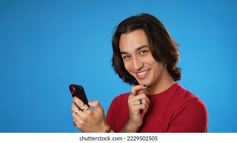 Happy smiling Hispanic Latino gender fluid young man 20s in red shirt using mobile cell phone online shopping, smiling isolated on solid blue background studio portrait - Powered by Shutterstock