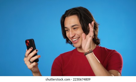 Happy smiling Hispanic Latino gender fluid young man 20s in red shirt talking waving in video chat call using mobile cell phone isolated on solid blue background studio portrait - Powered by Shutterstock