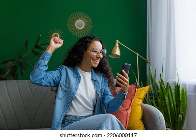 Happy And Smiling Hispanic Woman At Home Is Happy And Smiling Reading Online Message From Smartphone, Holding Hand Up Victory Gesture, Celebrating Triumph And Success.