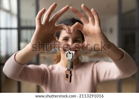 Happy smiling homeowner woman pose in living room hold keys from new first own house while makes heart love symbol with joined fingers. Accommodations apartment for newly-weds, dating services concept