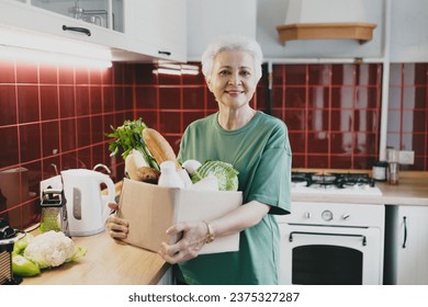 Happy smiling housewife holding carton box with fresh food products and vegetables for cooking dinner after returning from local market or grocery store, standing at kitchen. Home delivery service - Powered by Shutterstock