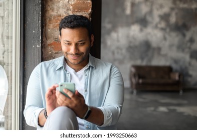 Happy and smiling indian dark-haired man in casual shirt using smartphone in loft style apartment, sits on windowsill texting, chatting online, web surfing, using new mobile app in rested atmosphere