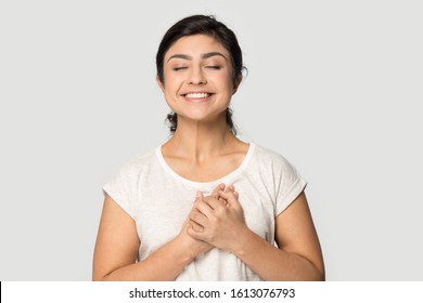 Happy smiling indian ethnicity millennial girl standing with closed eyes and folded hands on chest near heart, feeling thankful, dreaming about good future, luck isolated on grey studio background.