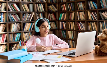 Happy Smiling Indian Junior Student Wearing Headphones Having Online Virtual Learning Language Class On Laptop. Cute Latin Schoolgirl Writing In Workbook Watching Video Meeting With Teacher.