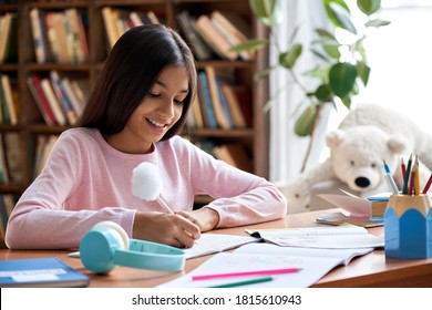 Happy smiling indian latin preteen school girl pupil studying at home sitting at desk. Smart cute hispanic kid primary school student writing in exercise book doing homework, learning at table.