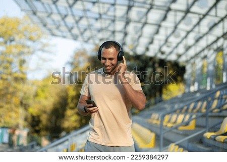 Happy and smiling Indian man in park stadium walking after active jogging and fitness classes, using phone and application for listening to online radio podcasts and music, sportsman in headphones.