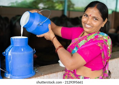 Happy smiling Indian woman busy working by pouring milk into container while looking at camera - concept of milk production, agri business, growth and empowerment.
