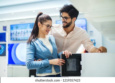 Happy Smiling Just Married Couple Shopping New Stuff For Their Home In Electronic Store. Choosing Together And Having Fun.