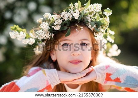 Happy smiling kid girl 6-7 year old wear knit colorful sweater and floral wreath hairstyle with flowers in park over nature background. Springtime. Spring season. Childhood. 