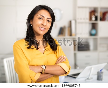 Happy smiling Latin American businesswoman standing in office with arms crossed looking at camera