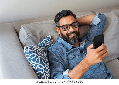 Happy smiling latin man using smartphone device while sitting on sofa at home. Mature indian man lying on couch reading messages on mobile phone. Hispanic guy with eyeglasses relaxing at home.