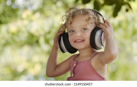 Happy Smiling Little Baby Girl Listening To Music Wearing Headphones, Close-up Portrait Of Funny Kid With Blue Eyes And Blond Curly Hair, Laughing Child Looking At Camera, Outdoor In Sunny Summer Day.