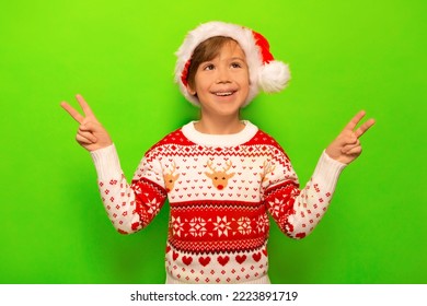 Happy smiling little boy in Santa Claus hat, Christmas sweater with deer and snowflakes pattern showing peace gesture or v sign with two fingers and looking up on a green background.