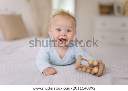 a happy smiling little cute 9 month old toddler baby boy in blue bodysuit trying to crawl and playing with a wooden rattle at home on the bed. eco friendly toys and motherhood concept