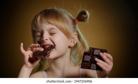 Happy Smiling Little Girl Eating Milk Chocolate Bar Isolated On Dark Background. Satisfied Face Of Pretty Caucasian Child Enjoying Unhealthy Sweet Food Indoors. Joyful Kid Eating Dessert Sweet Candies