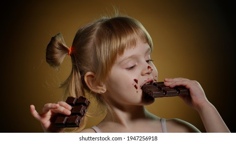 Happy Smiling Little Girl Eating Milk Chocolate Bar Isolated On Dark Background. Satisfied Face Of Pretty Caucasian Child Enjoying Unhealthy Sweet Food Indoors. Joyful Kid Eating Dessert Sweet Candies