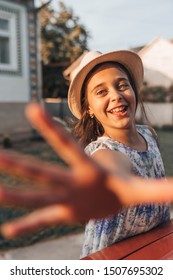 Happy Smiling Little Girl With Healthy Skinin Dress And White Hat Looking At The Camera And Reaching For The Camera, Summer Time, Outdoor Photo