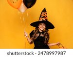 Happy smiling little girl wearing a witch costume holds pumpkin pot and holiday air balloons, celebrates Halloween on yellow background. Party for children, space for text. Kids trick or treating