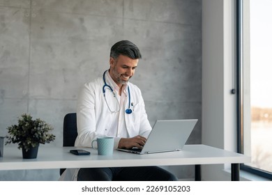 Happy smiling male doctor medical expert wearing lab coat using laptop computer at work in hospital, checking electronic files about patients, filling medical forms, having online virtual consultation - Powered by Shutterstock