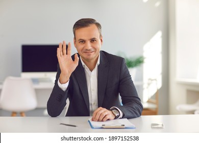 Happy Smiling Man In Suit Sitting At Office Desk, Looking At Camera And Waving Hand Saying Hello During Video Call. Webcam Portrait Of Business Coach, CEO, Financial Consultant Or University Professor