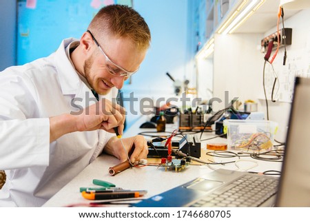 Happy, smiling man, technician, laboratory worker, testing and metering electronic cirquit board. Laptop in foreground.