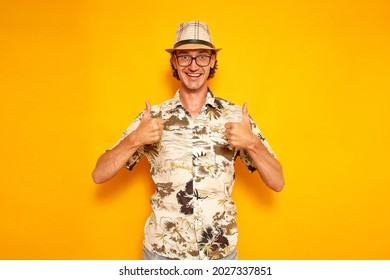 Happy Smiling Man Tourist Traveling Around World On Vacation Hawaiian Shirt Hat Glasses Rejoices Success Isolated Yellow Background Holds Thumbs Up Showing Everything Fine. Concept Recreation, People