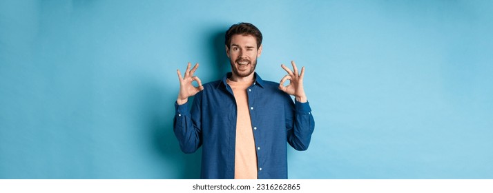 Happy smiling man winking and showing alright OK sign, say yes, approve and agree to something good, standing on blue background.