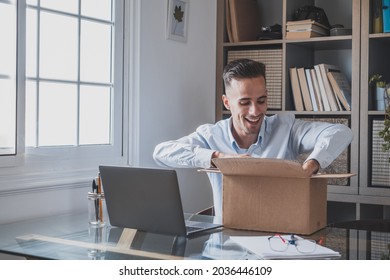 Happy Smiling Man Working And Opening A Small Box Just Delivered At Home Enjoying His Surprise Alone Having Fun. Young Caucasian Male Unboxing Some Present Or Gift At Office.