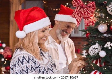 Happy Smiling Mature 50 Years Old Couple Wearing Santa Hats Decorating Christmas Tree At Home Together Celebrating Festive Merry Xmas Holiday Party Eve, Laughing, Having Fun, Feeling Joy.