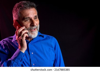 Happy Smiling Mature Bearded Man Talking On Mobile Isolated On Gel, Neon Light, Dark Background. Smiling Mature Guy Talking On A Mobile Phone Isolated On Neon Light Background.