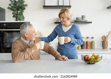 Happy and smiling mature married family drinking coffee on kitchen. Elderly couple spending weekend day at home, talking in bright room with modern interior, enjoying moment and beverage - Powered by Shutterstock