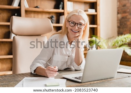 Happy smiling mature middle-aged businesswoman tutor boss ceo freelancer teacher doing paperwork, signing contracts, documents while working on laptop in office