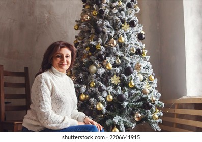 Happy Smiling Mature Woman Near Beautiful Xmas Tree At Home. Portrait Of Positive Cheerful Senior Female In White Knitted Sweater Sitting In Room Decorated For Christmas Or New Year