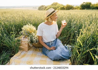 Happy Smiling Mature Woman Relaxing In Picnic In Countryside. Blond Lady In A Wheat Field. Slim Female With Basket Wild Flowers.