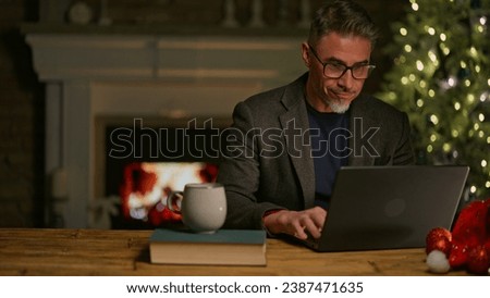 Happy smiling mid adult man using laptop computer at home, sitting at desk in Christmas decorated cosy room. Fireplace and Christmas tree in background. Businessman shopping online searching for gift.