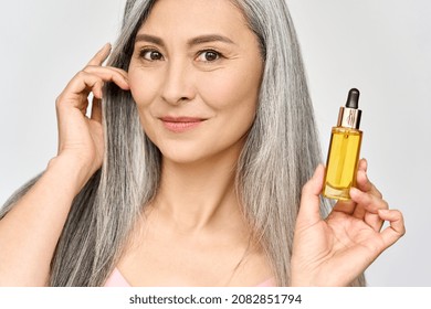 Happy Smiling Mid Age Asian Attractive Woman With Grey Silky Shiny Volume Hair Holding Bottle With Serum Essence Rich Nourishing Oil For Skincare And Haircare. Cosmetics Hair Face Treatment Concept.