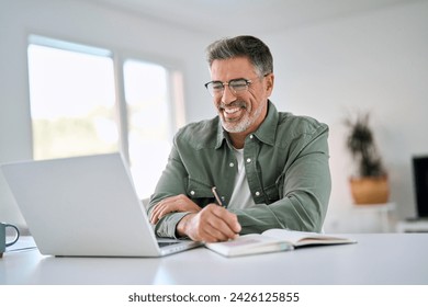 Happy smiling middle aged older mature man wearing glasses looking at laptop using computer writing notes watching webinar sitting at home table. Hybrid work, elearning, online learning concept. - Powered by Shutterstock